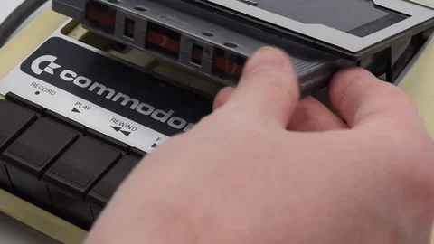 A hand puts a tape cassette in Commodore 64 vintage console to play a game. Stock-Footage 79854352