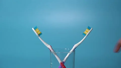 Hand puts two toothbrushes into a glass beaker on a blue background Stock Footage 145082441