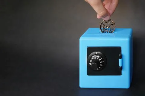 A hand is putting a clock-shaped coin in a blue safe-shaped piggy bank. Foto stock