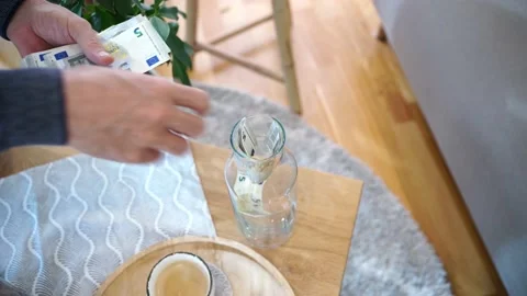 Hand putting five euro notes into a glass bottle in a living room. Saving money. Stock Footage 222899896