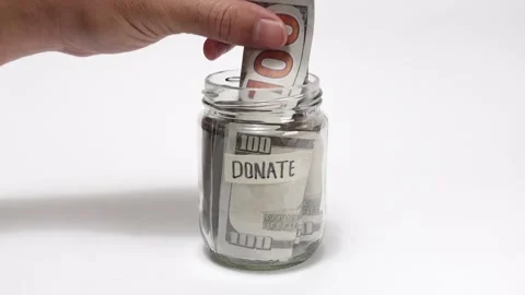 A hand putting a hundred dollar bill into a glass jar labeled 'Donate' Stock Footage 317108247
