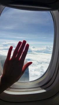 Hand raised in front of an airplane window during flight above a cloud layer. Pa Stock Photos