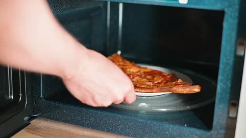 A hand reaches into a microwave to place a slice of leftover pizza on a plate. Stock Footage 338290176