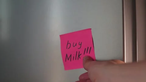 A hand reaches for a pink sticky note on a refrigerator door. The note has a Stock Footage 339096506