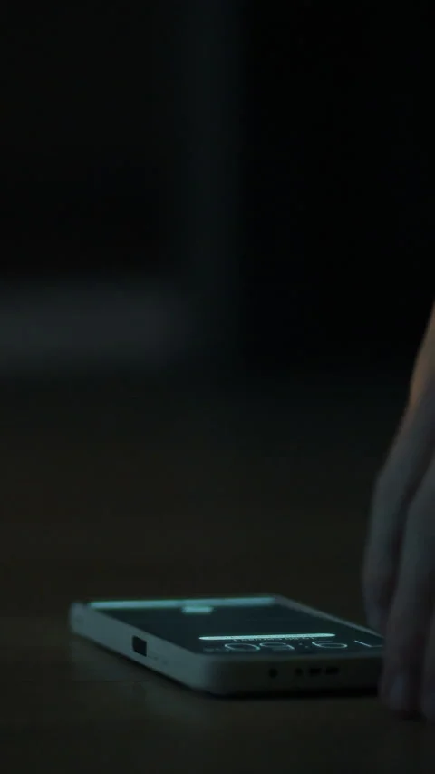 Hand reaching for smartphone on the floor beside a dark slipper in dim light Stock Footage 331596560