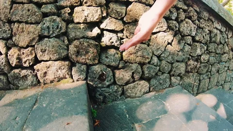 Hand reaching toward rough stacked volcanic stone wall beside weathered green Stock Footage 338440802