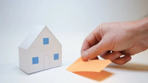 Hand rests next to a paper house purchase home symbol an orange post it Stock Footage 99722158