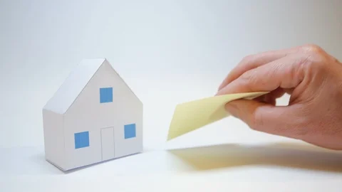 Hand rests next to a small paper house purchase symbol a yellow post it Stock Footage 99722506