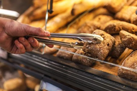 Hand Selecting Crusty Bread Rolls with Tongs at Bakery Display Stock Photos