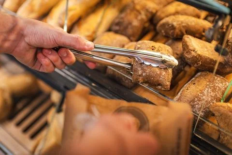 Hand Selecting Fresh Bread Rolls with Tongs at Bakery Display Stock Photos
