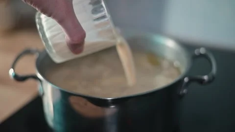 A hand is shown pouring liquid from a container into a pot of boiling soup on a Stock-Footage 338301400