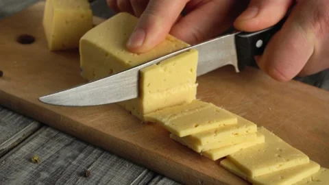 Hand slicing block of yellow cheese into even pieces on wooden cutting board Video stock 337831110