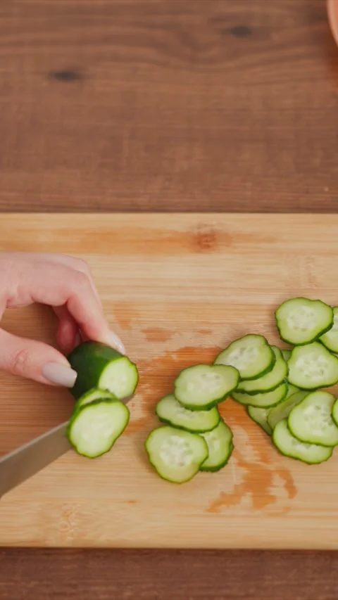 Hand slicing cucumber into thin rounds on wooden cutting board with increasing Stock Footage 346776310