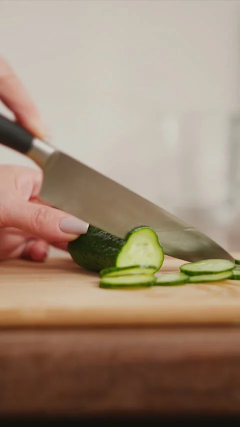 Hand slicing cucumber into thin rounds on wooden cutting board with knife Stock Footage 348034475