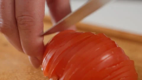 Hand slicing ripe tomato into thin even pieces on wooden board under warm Stock Footage 345657147