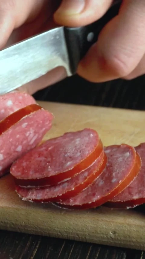 Hand slicing sausage into thin rounds on a wooden cutting board with a sharp Stock Footage 338395879