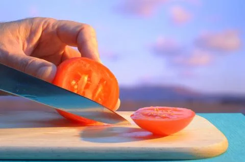 Hand slicing a tomato with a knife on cutting board on an island with pink cl Stock Photos