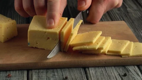 Hand slicing yellow cheese block into thin pieces on wooden cutting board with Stock Footage 337324052