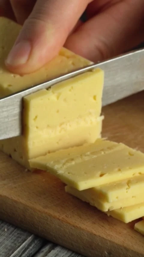 Hand slicing yellow cheese into even pieces on a wooden cutting board with a Video stock 338306671