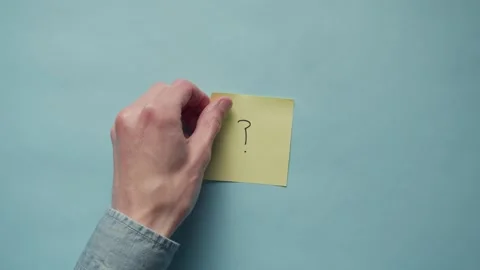 Hand slowly sticks a yellow post it note with question mark onto a blue backdrop Stock Footage 320557199