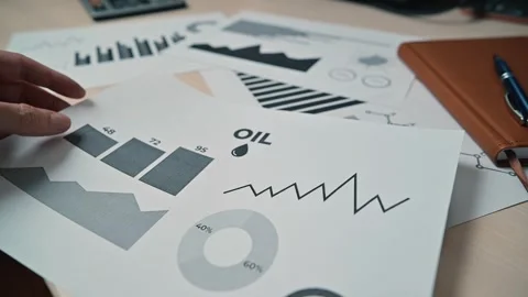 A hand sorts through printed graphs and charts related to oil market trends 库存影片 338301448