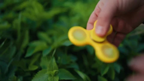 Hand Spinning a Bright Yellow Fidget Spinner Surrounded by Lush Green Leaves in Stock Footage 308335286