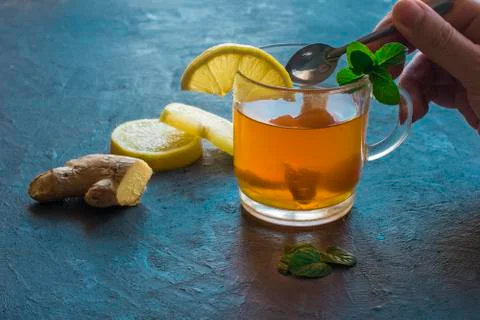 A hand with a spoon,a cup with ginger tea and lemon on a rustic blue table. Stock Photos