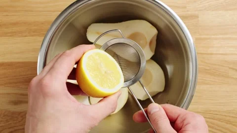 Hand squeezes lemon juice through sieve over pear halves in bowl. Video stock 344769722