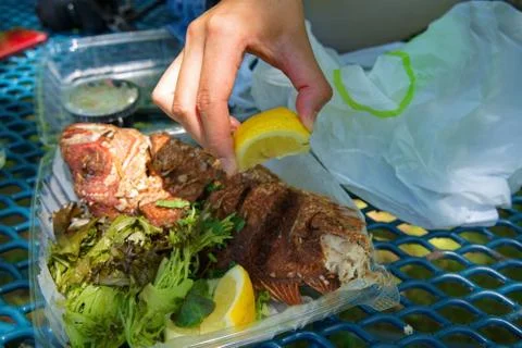 Hand squeezing a freshly cut lemon over a juicy take out fried red snapper. Stock Photos