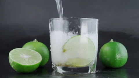 Hand squeezing lime into a glass with ice and lemon slices on a black background Stock Footage 220328037