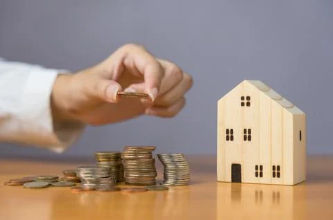 Hand stacking coins next to small wooden house model on wooden desk Stock Photos