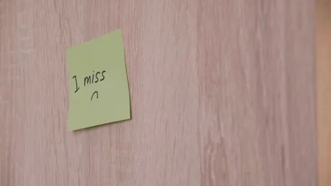 Hand sticking a yellow sticker with the text "I miss" on a light wood wall. Stock Footage 335506255