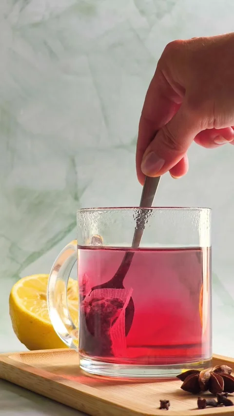 Hand stirring red herbal tea with spoon in clear glass cup, hot steam Video stock 336953630