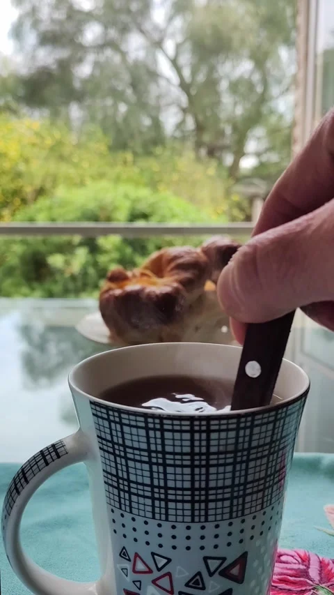 Hand stirring a steaming cup of coffee. A plate with a croissant is in the .. Stock Footage 286763342