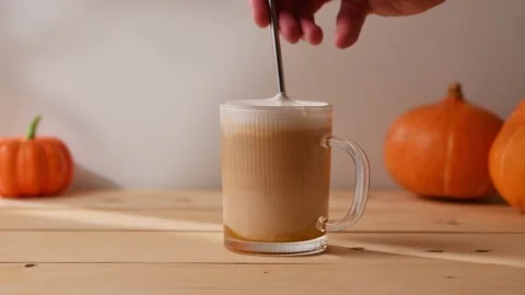 Hand stirs iced pumpkin latte in clear glass mug, blending rich pumpkin, milk, Stock Footage 313537804