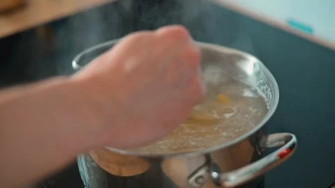 A hand stirs pasta in a pot of boiling water on a stove. The steam rises while a Stock Footage 338290448