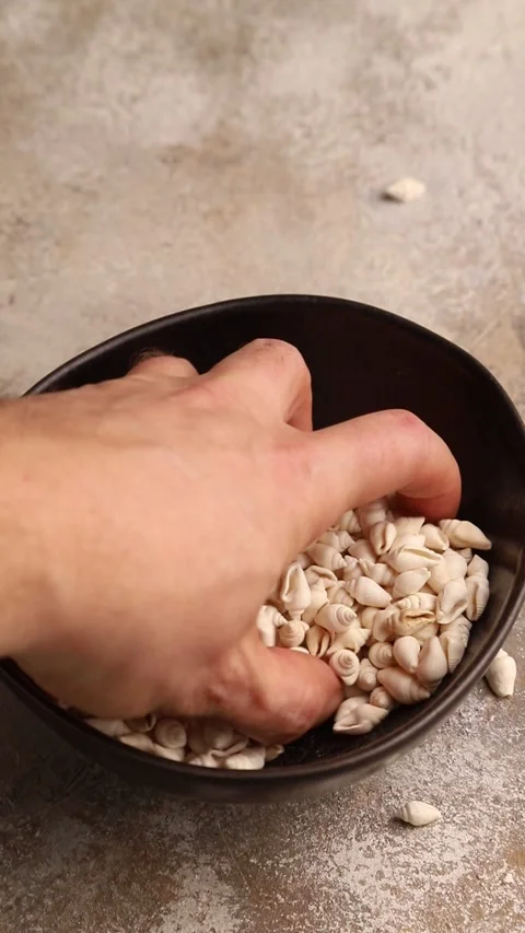 Hand stirs small white seashells pour into a ceramic bowl on a brown concrete Video stock 221546290