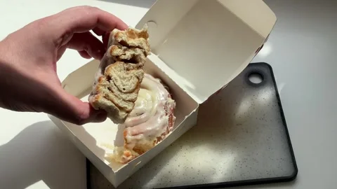 Hand taking half of sweet cinnamon roll bun with cream glaze Stock Footage 343981502
