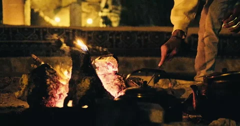 Hand tending glowing campfire logs in rustic stone fire pit at night Stock Footage 342729725