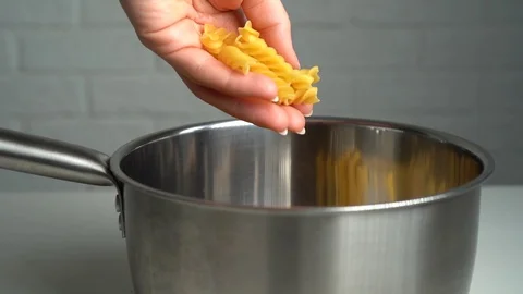 Hand throwing fusilli pasta into metal pot, white brick wall background. Stock Footage
