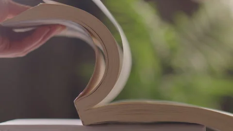 Hand Turning Thick Pages of Open Book with Blurred Green Ferns Background Vidéo 335474533