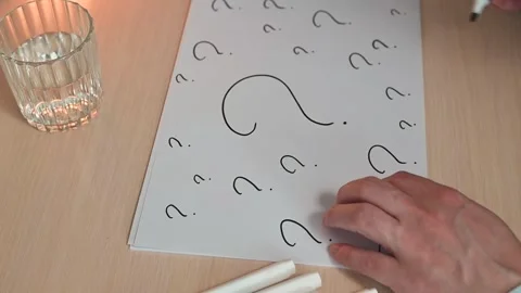 A hand uses a black marker to draw a question mark on a sheet of paper covered Stock-Footage 337558665
