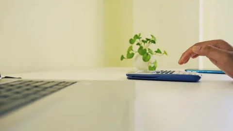 Hand using a calculator on a white desk with a laptop and a small plant Stock Footage 317737067