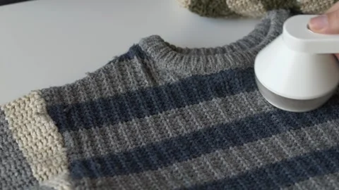 Hand using fabric shaver to remove lint from striped knit sweater on table Stock Footage 331010495