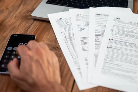 Hand using a smartphone calculator next to various tax forms on a wooden desk Stock Photos