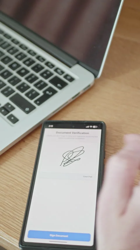 Hand using a smartphone to sign an e-document on a wooden desk next to a laptop. Vídeo Stock 335526722
