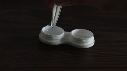 Hand using special tweezers to pick up contact lens from white lens case on dark Stock Footage 309309391