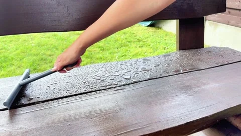 Hand Using Squeegee to Wipe Water Off Wet Outdoor Wooden Bench Surface 스톡 동영상 346499539
