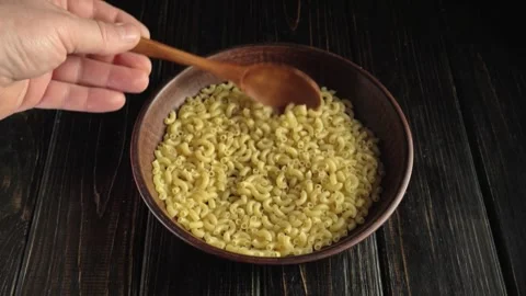 Hand using wooden spoon to stir cooked macaroni pasta in a brown bowl on a dark Stock-Footage 339030586