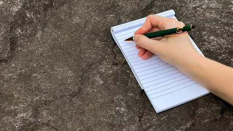 A hand writes in a notebook with a green pen on a stone surface Stock Photos